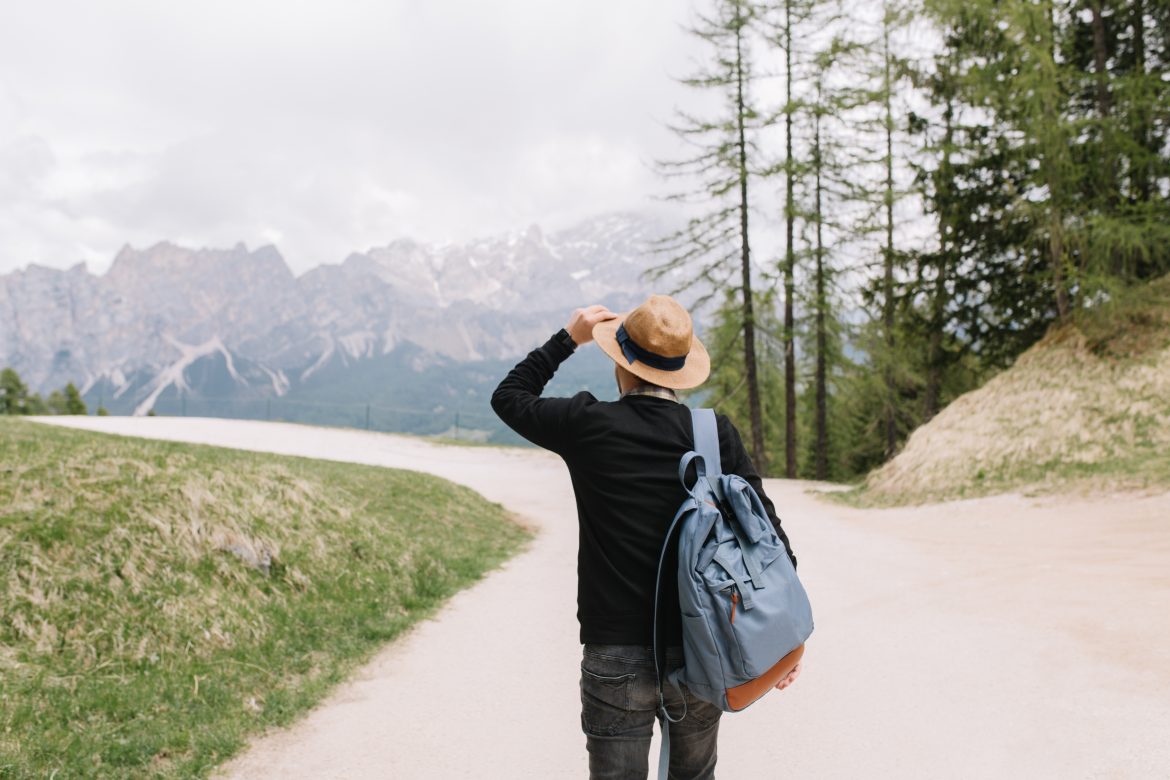 Stylish guy wearing hat admires the mountain scenery spending time outside in spring vacation. Outdoor portrait of male traveler with trendy backpack walking on the road near the green forest..